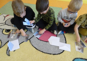 Trójka dzieci siedzi na dywanie, każde dziecko układa obrazek z części przedstawiający dinozaura.